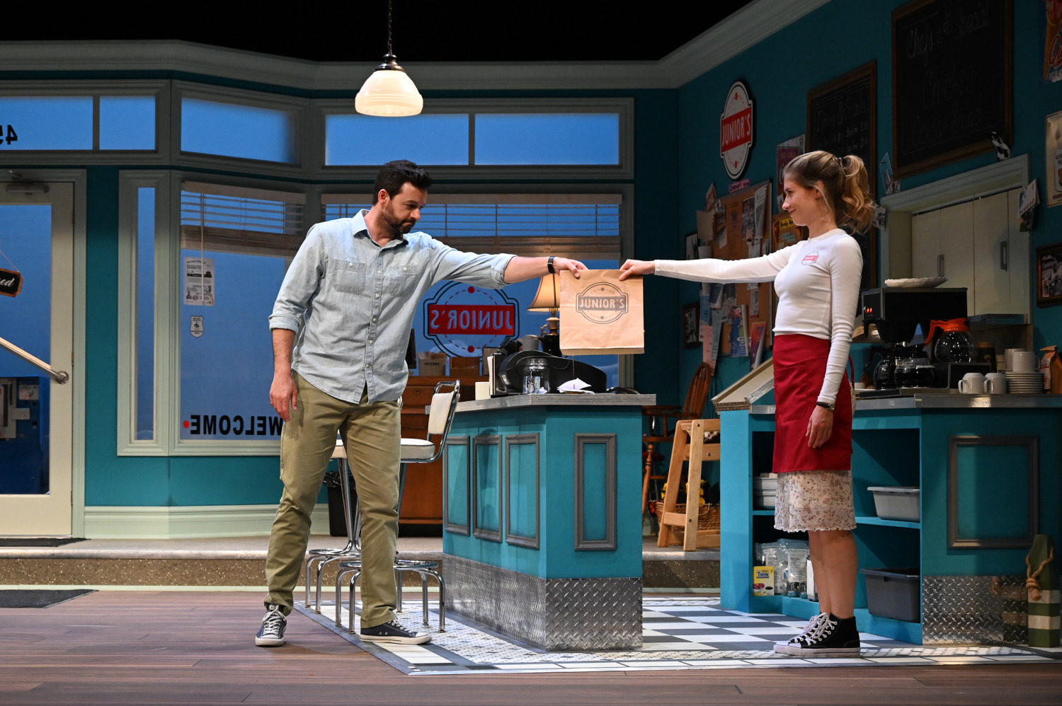 Two actors in a diner set, one passing a takeout bag to the other. The diner features a blue and white color scheme with a sign that reads 'Junior's'.