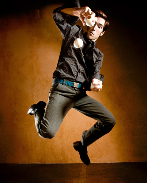 A male juggler mid-air, dressed in black with a blue belt, skillfully juggling three white balls against a warm-toned background. His dynamic pose and dramatic lighting enhance the theatrical energy.