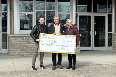 Three people pose outdoors in front of a building, holding an oversized check. The check, made out to Kirk Wallis, is for $5,175.00 from the 