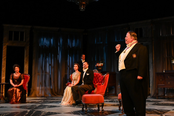 In this image from An Inspector Calls, a group of well-dressed characters is seated in an opulent, dimly lit drawing room. A man in formal attire, standing at the front, gestures passionately, while others sit attentively on plush, red velvet chairs. The atmosphere feels tense and dramatic, as the backdrop of long, flowing curtains and ornate furniture sets a mood of mystery and suspense. The actors' period costumes, particularly the men in tuxedos and the women in elegant gowns, reflect the early 20th-century setting of the play.