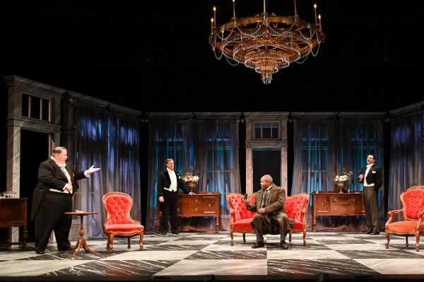 In this image from An Inspector Calls, four male characters are positioned around an elegant drawing room. A large, ornate chandelier hangs above, illuminating the scene. One man in formal evening attire stands to the left, gesturing towards a man seated on a red velvet chair at center stage, who appears engaged in conversation. Two other men in tuxedos stand at the back, observing the interaction. The room is filled with vintage furniture, including red velvet chairs, contributing to the atmosphere of formality and tension.