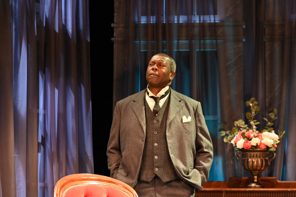 In this image, a male actor stands confidently in a well-tailored three-piece suit, embodying a character of authority and dignity. He wears a grey vest and jacket, paired with a white pocket square, and a loosely tied black scarf. The backdrop consists of elegant drapes and soft lighting, creating an air of sophistication and formality. A red velvet chair is partially visible in the foreground, and a vase of colorful flowers sits atop a wooden table behind him, adding to the refined setting of the scene.