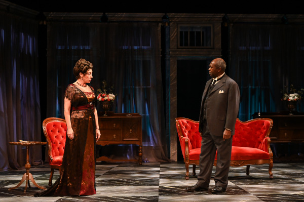 In this image from An Inspector Calls, two characters stand facing each other in an intense moment. The woman, dressed in a rich, dark, lace-trimmed gown with red accents, gazes sternly at the man, who is wearing a formal grey suit with a vest and tie. They stand on opposite sides of the stage, separated by red velvet chairs and a small side table. The elegant, dimly lit setting, with ornate furnishings and draped curtains, reinforces the tension and formality of the scene.