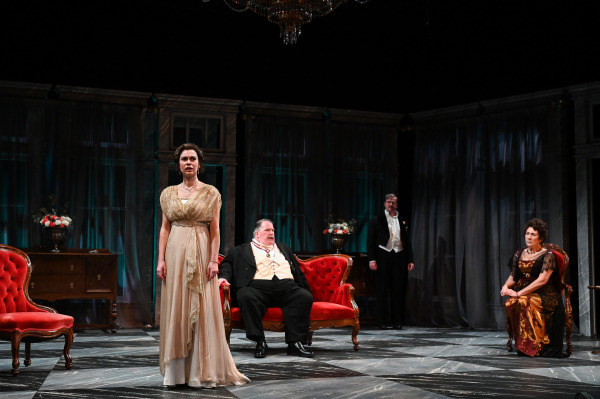 In this image from An Inspector Calls, a woman in an elegant cream-colored gown stands at the center, looking outward with a solemn expression. Behind her, a man in a tuxedo sits in a red velvet chair, while two others, also dressed in formal evening wear, stand and sit at a distance, contributing to a mood of tension. The stage is set with ornate furnishings, including rich red chairs, dark curtains, and soft lighting, enhancing the dramatic atmosphere of the scene.