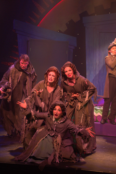 A group of four characters dressed in tattered, ragged clothing with distressed facial expressions huddle together on stage. They appear to be pleading or lamenting, with hands extended and mouths open in expressive poses. The lighting casts a warm glow on their faces, highlighting their emotional intensity. The backdrop features large, dark geometric shapes with a subtle sparkle effect, adding depth to the scene. Another character in a suit can be seen standing in the background, slightly out of focus, suggesting a contrasting role to the group in rags.