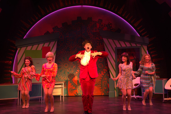 A lively stage scene featuring a central character dressed in a bright red suit with a ruffled white shirt, passionately gesturing with their hands on their chest and singing or shouting. Surrounding them are four other characters in vibrant, patterned 1960s-style dresses, each striking dynamic poses and smiling. The backdrop has a large circular design with colorful, abstract patterns and gear shapes, creating a retro, energetic atmosphere. The actors appear to be mid-dance, capturing the high energy and fun spirit of the performance.