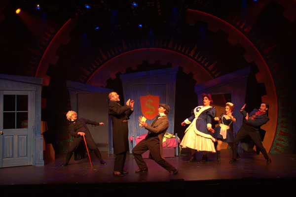 A comedic stage scene with six characters in exaggerated poses, all seemingly caught in a moment of surprise or shock. On the left, an older character with wild white hair and a cane leans forward, gesturing dramatically. The center of the stage features a character in dark formal attire recoiling, while another character in a cap and vest lunges towards them, hands raised in a pleading or urgent manner. To the right, two women in blue and white maid and governess costumes, one of whom has a large hoop skirt, react with exaggerated expressions of alarm. The background features a vibrant set with large cog-like designs and a shield emblem, adding to the playful, animated atmosphere of the scene.