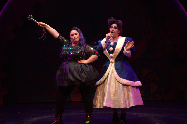 Two characters stand side by side on stage in an energetic, lighthearted pose. On the left, a performer with purple hair, dressed in a sparkly black top and black skirt, holds a wand aloft with a triumphant expression. On the right, a performer in a blue and white fur-trimmed dress with puffy sleeves and a voluminous skirt sings into a microphone, exuding dramatic flair. The background features large cogwheel designs, adding a whimsical, industrial feel to the scene. Both characters display exaggerated expressions, capturing the comedic and playful atmosphere of the performance.