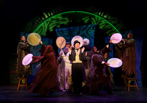  The image shows a theatrical scene featuring a central character dressed in a formal black suit, a gold vest, and a top hat, appearing smug and confident as they stand amidst a group of performers. Surrounding the central figure are actors in tattered, dark costumes resembling beggars or ghosts, energetically holding oversized coins with Canadian currency designs. The background is a vibrant green, with glowing arch-like structures and stage props resembling wooden doors. The dramatic lighting highlights the central figure and the coins, creating a whimsical and satirical atmosphere that blends themes of wealth and poverty.