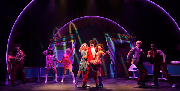 The image depicts a lively musical theatre scene set against a colorful, retro-inspired backdrop. A group of performers is positioned across the stage, dressed in vibrant 1960s-style costumes, including patterned dresses, flared pants, and knee-high boots. The central figure, wearing a bold red suit with a ruffled white shirt, is flanked by two female performers in brightly colored dresses, all striking playful poses. The stage is illuminated with vibrant purples, greens, and yellows, enhancing the festive atmosphere. The background features a decorative arch adorned with garlands, clock motifs, and a colorful design reminiscent of a psychedelic era, creating a whimsical and dynamic setting.