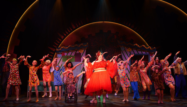 A vibrant musical theatre performance featuring a large ensemble cast dressed in colorful, retro-inspired costumes. At the center of the scene stands a performer in a dramatic bright red dress adorned with sparkling embellishments and a festive headdress, radiating confidence and joy. Surrounding them are performers of diverse ages and appearances, dressed in 1960s and 1970s-style floral and patterned outfits, including bell-bottoms, mini dresses, and headbands. Some performers use mobility aids, emphasizing inclusivity in the cast. The backdrop is a whimsical design with gears, clocks, and garlands, illuminated in warm tones, creating a lively and celebratory atmosphere. The cast strikes synchronized, playful poses, exuding energy and unity.