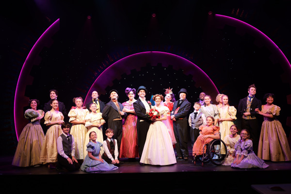  A full-cast photo from a musical theatre production featuring performers in elegant period costumes. At the center, a couple dressed in wedding attire—a groom in a top hat and tuxedo and a bride in a white gown holding a bouquet of red flowers—stand smiling and holding hands. Surrounding them, the ensemble cast is arranged in two rows, with children and some seated performers in the front and adults in the back. The costumes include ball gowns in pastel shades, suits, and formal wear. The backdrop features a glowing arch in purple hues, with starry lights adding to the festive and celebratory atmosphere. The composition radiates joy, unity, and grandeur.