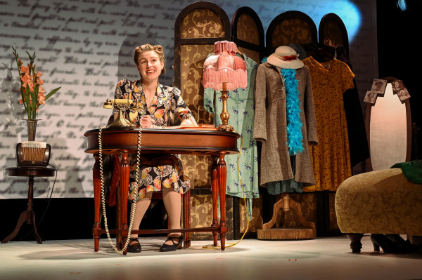 In this image, a female performer sits at a wooden desk, smiling as she engages with an antique rotary telephone. She wears a vintage floral dress in muted tones, with curled hair styled in a 1940s fashion. The stage is set in a cozy, intimate atmosphere with a screen divider in the background adorned with vintage coats, hats, and a bright blue feather boa. A pink lamp adds warmth to the scene, while an old-fashioned radio sits on a side table, enhancing the nostalgic mood of the production.