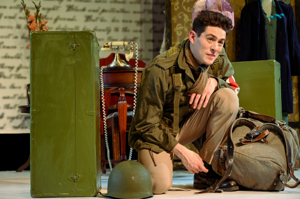 In this image, a male performer, dressed as a soldier in military gear, kneels on stage while adjusting his boot. He is wearing a green army jacket, scarf, and khaki pants. Beside him is a large green trunk, an army helmet, and a worn duffel bag, suggesting he's preparing for a journey or military mission. Behind him, part of a vintage telephone is visible on a wooden desk, adding a nostalgic touch. The backdrop features handwritten letters, reinforcing the emotional and historical themes of the scene.