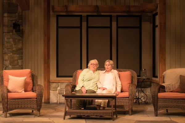 A scene from Doris and Ivy in the Home with two people sitting close together on a wicker sofa, looking thoughtful, with a rustic indoor set in the background.