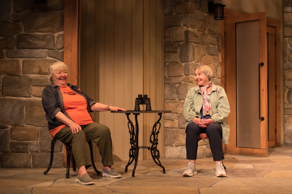 A scene from Doris and Ivy in the Home with two people sitting on a porch with stone walls, smiling and talking.