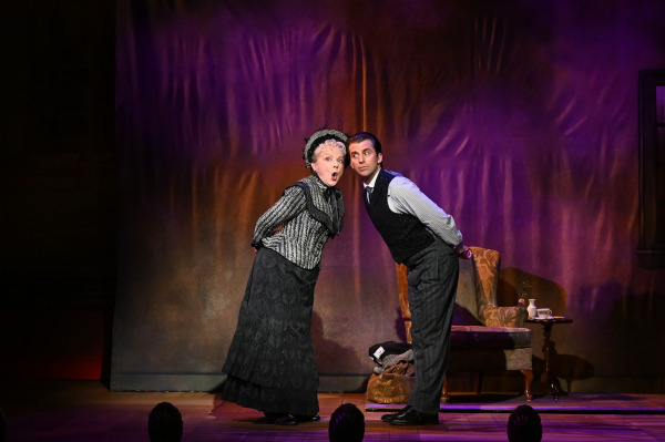 In this image from A Gentleman's Guide to Love and Murder, two actors are engaged in a comedic moment. The woman, dressed in a Victorian-era black and white ensemble with lace details and a bonnet, stands with a humorous expression, leaning forward. Beside her, a man in a vest and striped shirt mirrors her posture, both leaning toward the audience. The background is dimly lit with purple and warm tones, enhancing the playful and theatrical mood of the scene. A small side table with a teacup is visible in the background.