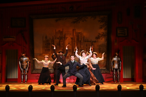 In this image from A Gentleman's Guide to Love and Murder, a group of actors is posed mid-dance in a lively, stylized formation. The performers, dressed in Edwardian-era costumes with long skirts and hats, stand in front of a grand backdrop of a large estate. The central actor, in a tailored suit, crouches with arms outstretched while the others mirror his raised-arm gestures. The set is adorned with suits of armor, giving the scene a grand, historical atmosphere, enhanced by the warm lighting on stage.