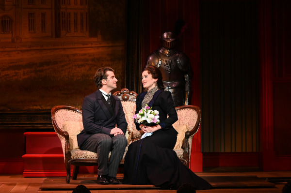 In this image from A Gentleman's Guide to Love and Murder, two characters are seated together on an elegant Victorian-style loveseat. The man, dressed in a dark suit, gazes lovingly at the woman beside him, who holds a bouquet of flowers and wears a dark, period-appropriate gown. Behind them, a suit of armor stands guard, adding a touch of grandeur and formality to the scene. The actors’ expressions and intimate positioning suggest a tender or emotional moment between the characters.