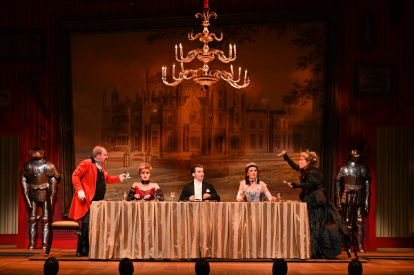 In this image from A Gentleman's Guide to Love and Murder, five characters are seated at a long dining table, with a grand castle painting as the backdrop. The man on the far left, dressed in a red coat, raises a glass as he interacts with a seated woman in a royal gown. The other characters look shocked or engaged in conversation, with one woman on the far right standing and gesturing dramatically. The set includes suits of armor, giving the scene an opulent, formal atmosphere, while the chandelier above adds to the grandeur.