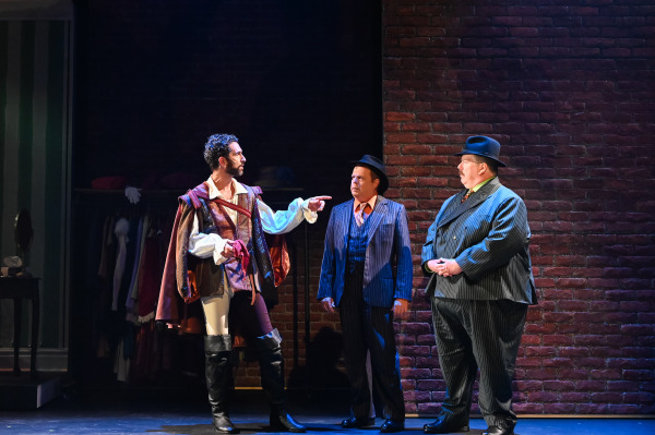 An intimate scene set against a dark brick wall. One performer is dressed in a rich Renaissance costume, complete with a cape and boots, and is pointing while speaking to two others dressed in 1940s-style suits and fedoras. The lighting is moody, highlighting the tension and seriousness of the moment, with the Renaissance garb contrasting against the modern, gangster-like suits.