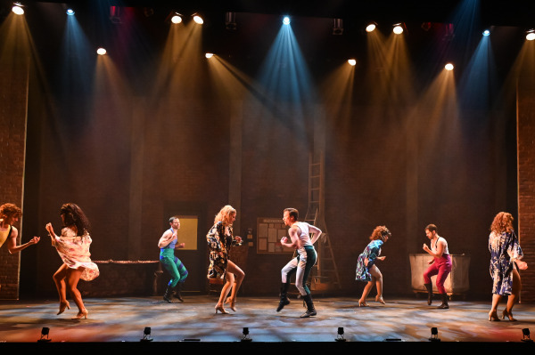 A high-energy dance number featuring several performers. The scene is set in a dimly lit room with brick walls and scattered props, including a ladder. The dancers, dressed in a mix of 1940s attire and Renaissance costumes, are engaged in lively movement, with bright spotlights highlighting their actions. The dynamic lighting and varied costumes create a sense of exuberance and theatrical flair.