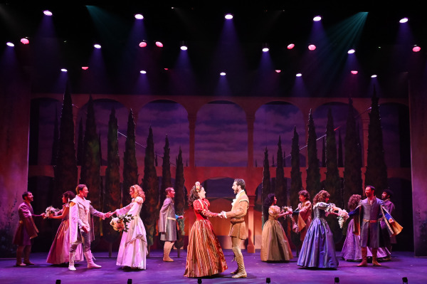 A romantic scene set in a picturesque outdoor courtyard with tall cypress trees and a dusky sky. Performers in Renaissance costumes dance and interact with each other in pairs, holding hands and smiling. The lighting is soft and warm, creating a magical atmosphere as they perform a choreographed dance. The scene evokes a sense of elegance and classic romanticism, characteristic of the golden age of theatre.