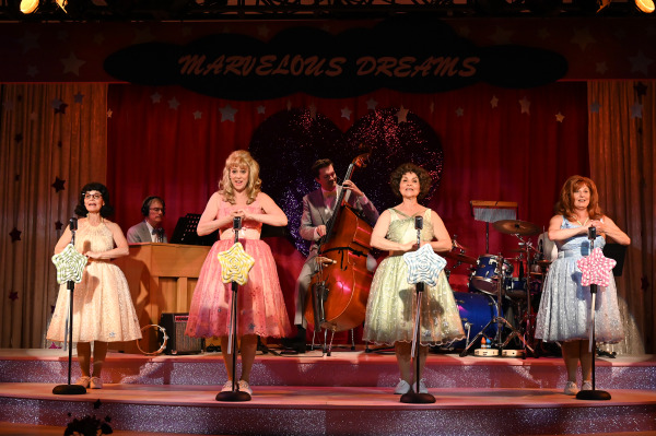 Four performers in glittery dresses sing on stage with vintage microphones. The backdrop reads 