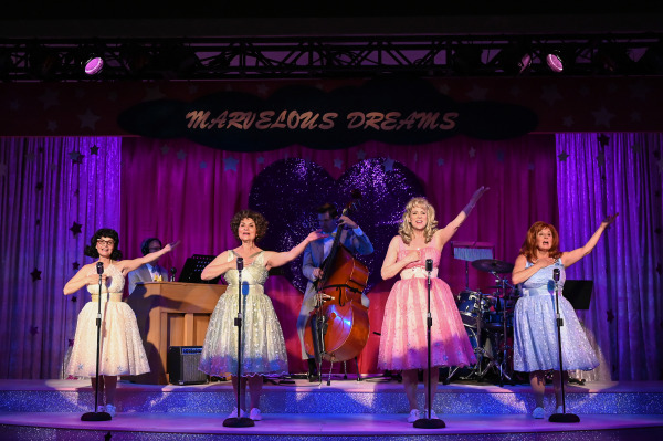 Four performers in glittery dresses sing on stage with vintage microphones. The backdrop reads 