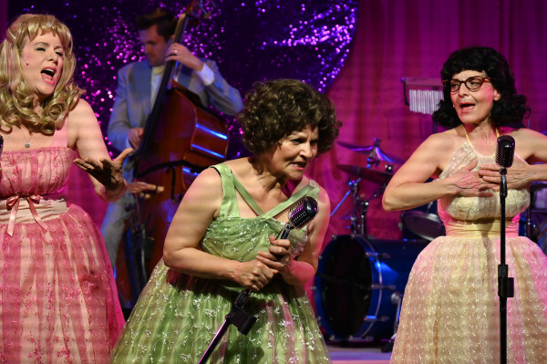 Three performers in colorful, 1950s-style dresses sing into vintage microphones on stage. The performer on the left wears a pink dress, the one in the center wears a green dress, and the one on the right wears a cream dress with glasses. They are passionately engaged in their performance, with a double bass player and a sparkling pink backdrop in the background, adding to the lively and nostalgic atmosphere of the scene.