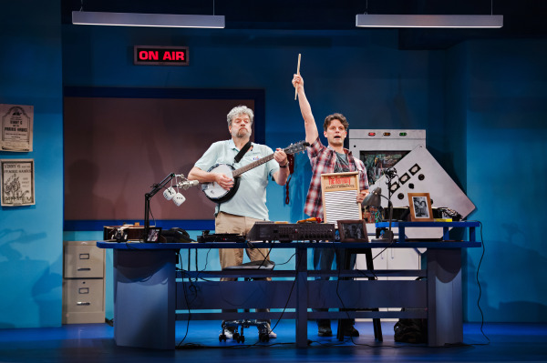 Two performers on stage in a radio studio set. One plays a banjo, while the other raises a drumstick and holds a washboard. The 'ON AIR' sign is lit above them, and the studio is filled with radio equipment and decorations, creating a lively scene from the play 