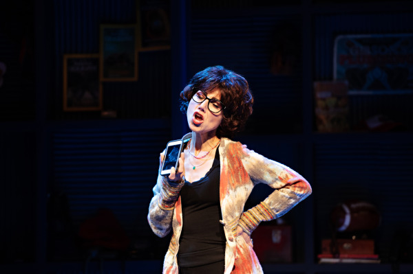 A performer is standing on stage, mid-sentence, holding a smartphone close to their face as if recording or using it for a voice call. The performer is dressed in a casual outfit, featuring a patterned cardigan with warm colors over a black top. They wear large, stylish glasses, and their hair is styled in loose curls. The expression is animated, suggesting they are in the midst of an impassioned or humorous moment. The background is dimly lit, with some details of a set that includes posters and various objects, creating a casual, radio-station-like atmosphere.