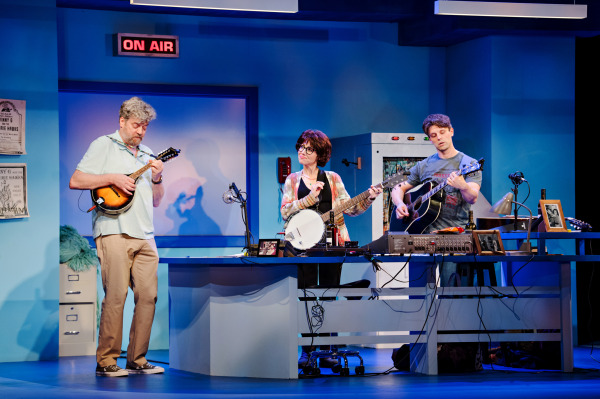 Three performers are engaged in a lively musical performance on stage. Each is playing an instrument: the performer on the left with a mandolin, the center performer with a banjo, and the performer on the right with an acoustic guitar. All three are singing passionately. The stage is set up like a radio station or recording studio, with equipment such as microphones, a soundboard, and various desk items, including a bottle of wine. The backdrop is a cool blue, giving a modern, light-hearted vibe to the scene. The performers' expressions and body language convey joy and enthusiasm as they perform together.