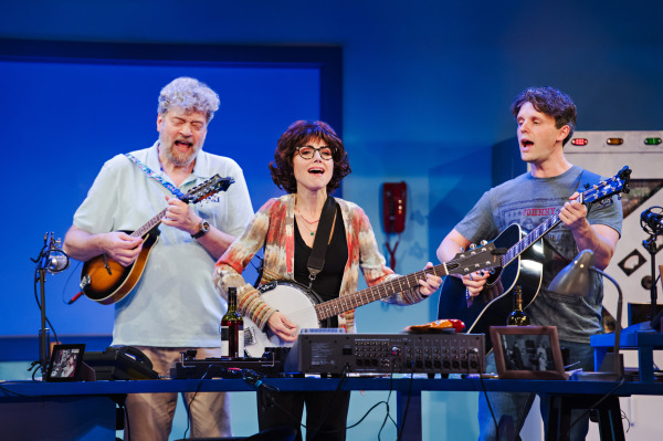 Three performers sing and play instruments on stage in a radio studio set. They are engaged and smiling, with the performer in the center playing a banjo, flanked by performers playing guitars. The studio set is filled with radio equipment, creating a vibrant musical moment from the play 