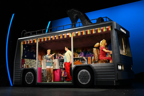Three performers in colorful costumes on stage in a bus set labeled 
