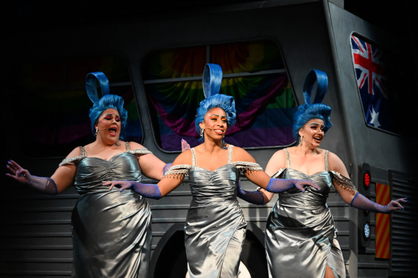 Three performers in glamorous silver dresses with blue gloves and towering blue wigs, sing and dance on stage. They are positioned in front of a bus set decorated with rainbow flags and an Australian flag. The scene captures a lively and vibrant moment from the musical Priscilla Queen of the Desert.