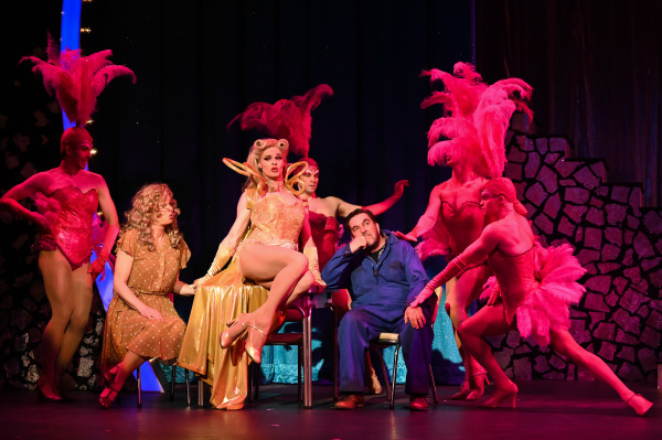 A scene from Priscilla Queen of the Desert featuring performers in elaborate costumes. In the center, a performer in a glamorous gold dress and matching gloves sits confidently with legs crossed, flanked by other performers in vibrant pink feathered outfits. Another performer in a brown polka-dot dress and a man in blue coveralls sit on either side, both engaged in the scene. The background is dark, making the colorful costumes and dynamic poses stand out dramatically.