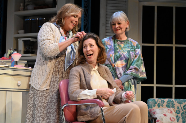 A scene from Steel Magnolias featuring three people in a beauty salon, one styling the hair of a seated person who is laughing, while another person stands beside them, smiling.
