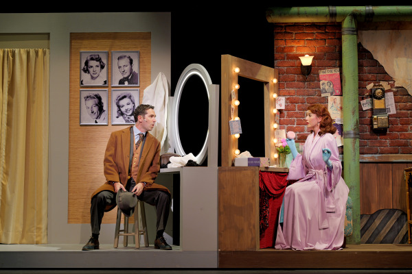 A theatre scene depicting a man and a woman in a dressing room. The man, dressed in a brown coat, tie, and hat, sits on a stool looking at the woman. The woman, dressed in a pink robe with gloves, sits at a vanity table with lit mirrors, holding a powder puff to her face. The background features vintage-style photos, a green pipe, and a brick wall adorned with posters and a rotary phone, evoking a nostalgic atmosphere.