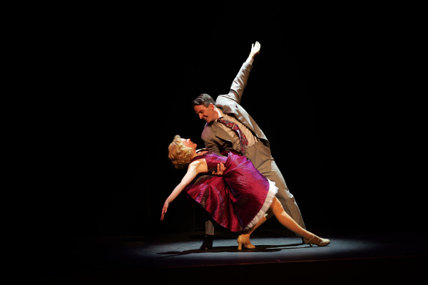 A dramatic dance moment on a dimly lit stage features a man and woman in an elegant pose. The man, dressed in a light grey suit and tie, leans into a deep dip, holding the woman securely as she arches backward. She wears a vibrant purple dress with a full skirt and beige heels, her blonde hair softly curled. The spotlight highlights their graceful posture against the black background, emphasizing the emotional intensity and precision of their performance.