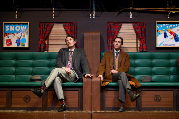 Two men sit on a vintage train's green tufted bench seat, framed by wooden paneling and plaid curtains. On the left, a man in a grey suit with a patterned tie leans back with a thoughtful expression, his hat resting on the seat beside him. On the right, another man wears a brown coat over a suit and tie, sitting upright and gazing forward with a composed expression. The scene is accented by retro-style ski-themed posters and a cozy, nostalgic atmosphere, suggesting a mid-20th-century train journey.