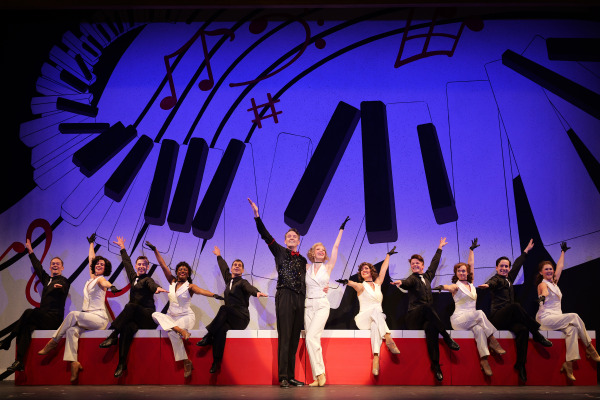 A lively ensemble performs against a vibrant backdrop of oversized piano keys and musical notes in black and red on a blue background. Center stage, two lead performers, one in a sparkling black vest and another in a glamorous white outfit, raise their arms triumphantly while smiling. Flanking them, the ensemble, dressed in elegant black and white costumes, sits on a red and white platform with one leg kicked high in perfect synchronization, exuding energy and joy in a classic musical theatre pose.