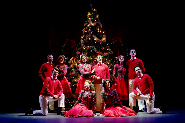  A group of performers dressed in festive red and plaid costumes pose in front of a beautifully decorated Christmas tree. The tree is adorned with twinkling lights, ornaments, and ribbons, creating a warm and cheerful holiday atmosphere. The cast, arranged in a mix of standing and kneeling positions, smiles warmly, radiating joy and holiday spirit. The stage lighting highlights the vibrant colors of their costumes and the glittering decorations on the tree, while the background remains dim, focusing attention on the ensemble.