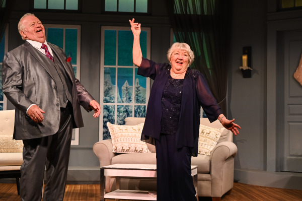 In this joyful scene from Merry and Bright, an older man and woman share a lighthearted moment in a cozy living room decorated for the holidays. The man, dressed in a shiny silver suit with a red pocket square, laughs heartily, while the woman, wearing a deep blue dress with a sheer jacket, raises her hand in a celebratory gesture, smiling widely. Behind them, large windows reveal a snowy winter landscape, enhancing the festive atmosphere. The room is warmly decorated with comfortable furniture and holiday-themed pillows.