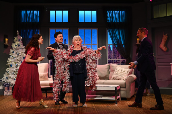 In this festive scene from Merry and Bright, an elderly woman stands in the centre, playfully wrapped in a long strand of red and white tinsel. She looks surprised and amused, with her arms raised in a dramatic pose. Surrounding her, a man in a suit and two other younger adults, one in a dark suit and the other in a red dress, smile and laugh as they celebrate together in a cozy, holiday-decorated living room. A beautifully decorated white Christmas tree adorned with blue ornaments and wrapped presents is visible in the background, adding to the festive ambiance of the scene.