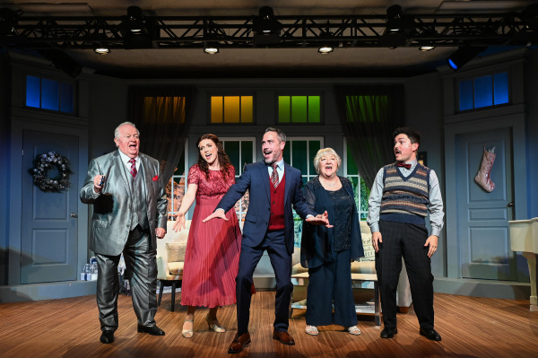 Five actors stand in a row on stage, mid-song, radiating energy and joy as they perform together. From left to right, a man in a shiny silver suit holds a remote with a jovial expression; a woman in a flowing red dress passionately sings; a man in a navy suit with a red vest gestures enthusiastically; an elderly woman in a dark outfit smiles widely, singing along; and a younger man in a sweater vest and bow tie looks to his left with a surprised expression. The stage is set with cozy holiday decor, including a wreath on the door and a Christmas stocking on the wall, capturing a festive and heartwarming moment.